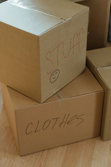 Three cardboard boxes stacked in a home interior, with the top box labeled 'STUFF' in red marker and a small smiley face drawn below the label, and the lower box labeled 'CLOTHES' in black marker. The boxes are placed on a wooden floor, with other similar boxes partially visible in the background, indicating an active packing or moving process. This scene reflects the organization involved in home relocation, with clear labels assisting in the efficient handling and unpacking of belongings. The natural lighting highlights the cardboard texture and the handwritten labels, aligning with the context of furniture transport and packing during a move managed by a professional removals service such as Man with Van Barnes.