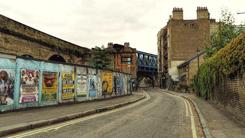 A narrow, gently curved street scene outside Barnes Bridge station featuring a weathered brick wall on the left, covered with colorful posters advertising events and concerts, and a brick building with small balconies on the right side, partially obscured by green foliage. A blue metal railway bridge spans across the top of the street, with a pedestrian underpass visible beneath it. The road is lined with double yellow lines, indicating parking restrictions, and appears to be in a residential or urban area undergoing a home relocation process. The lighting suggests daylight with an overcast sky, and the scene is typical of urban house removals or furniture transport activities near the station, with a focus on the local environment relevant to moving logistics and packing and moving services provided by Man with Van Barnes.