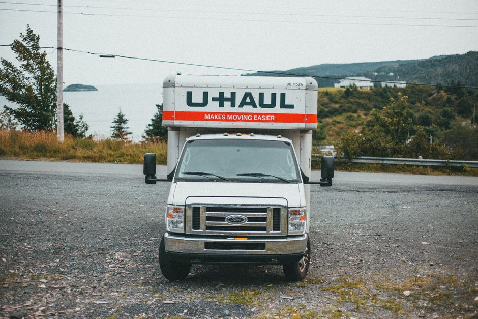 The image shows the rear of a white U-Haul moving truck parked on a gravel surface beside a rural road, with dense green trees and shrubbery in the background under an overcast sky. The truck's back d